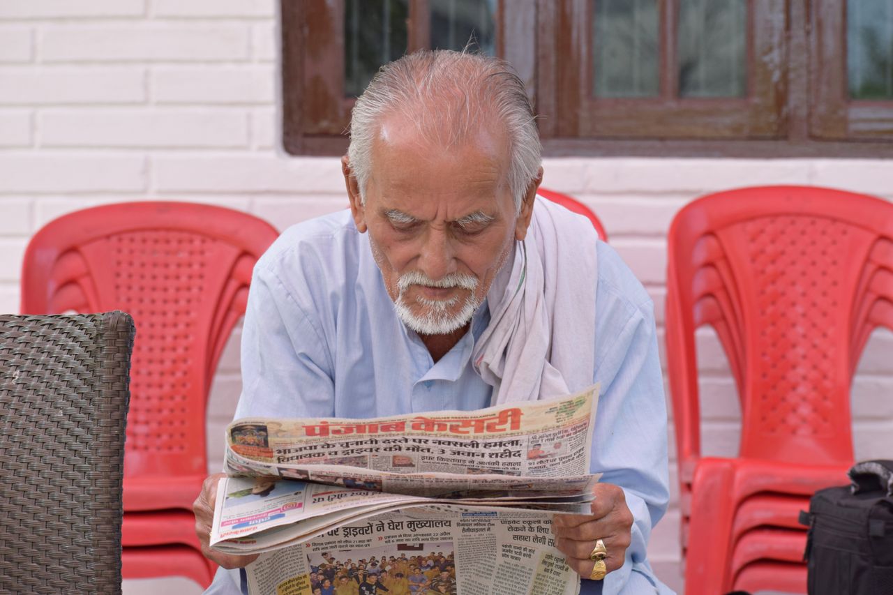 hero-img-01 Elderly man engrossed in reading a newspaper while seated outside with red chairs around.