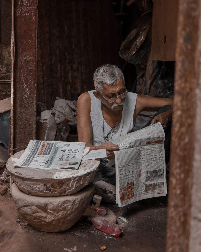 gallery-3 An elderly man sits reading a newspaper in a rustic indoor setting, surrounded by traditional items.