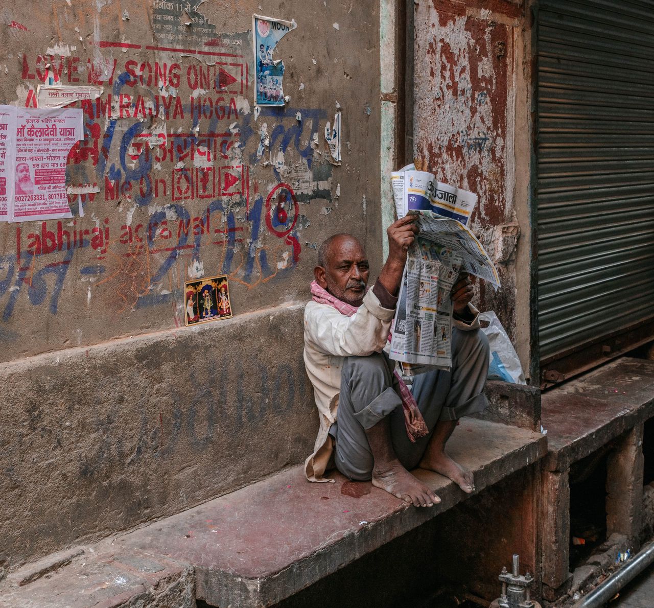 hero-img-02 An elderly man sitting outdoors on a street in Mathura, India, reading a newspaper.