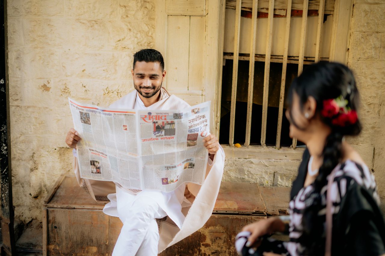 services-01 A man in traditional attire reading a newspaper outside with a woman visible in the foreground.