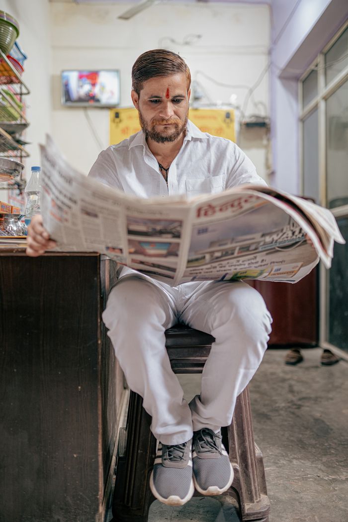 services-02 A man in traditional attire reads a newspaper in a shop in Varanasi, India, capturing local culture.