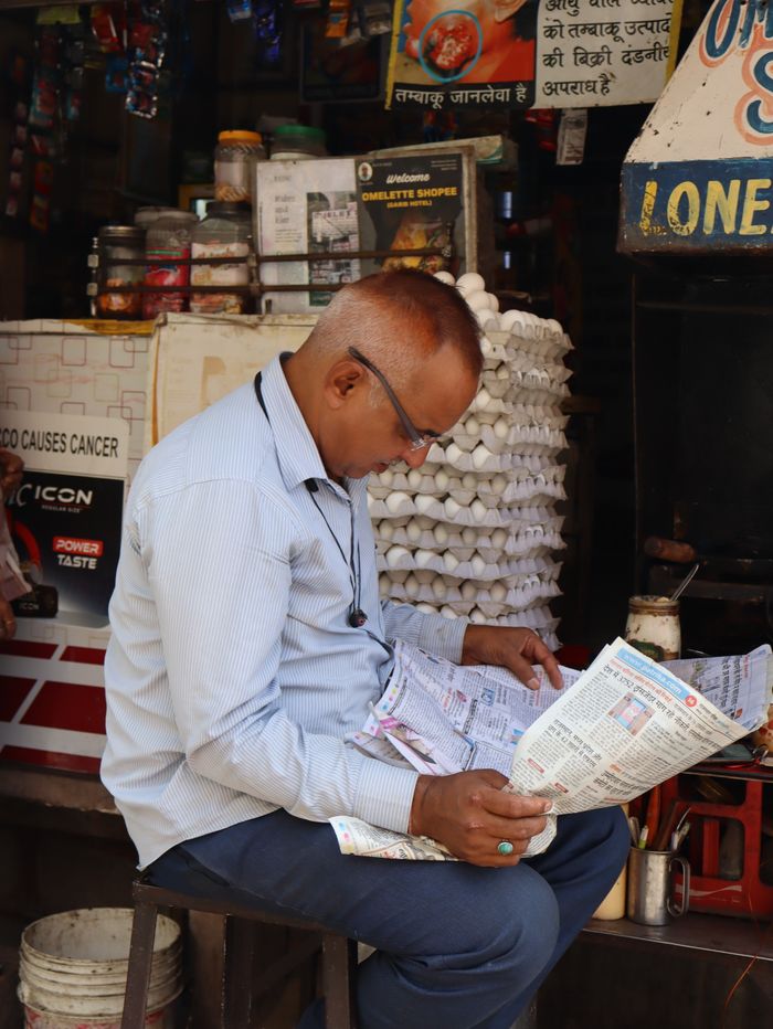 services-img Candid street portrait of a man engrossed in reading a newspaper at a bustling market stall in Jodhpur, India.