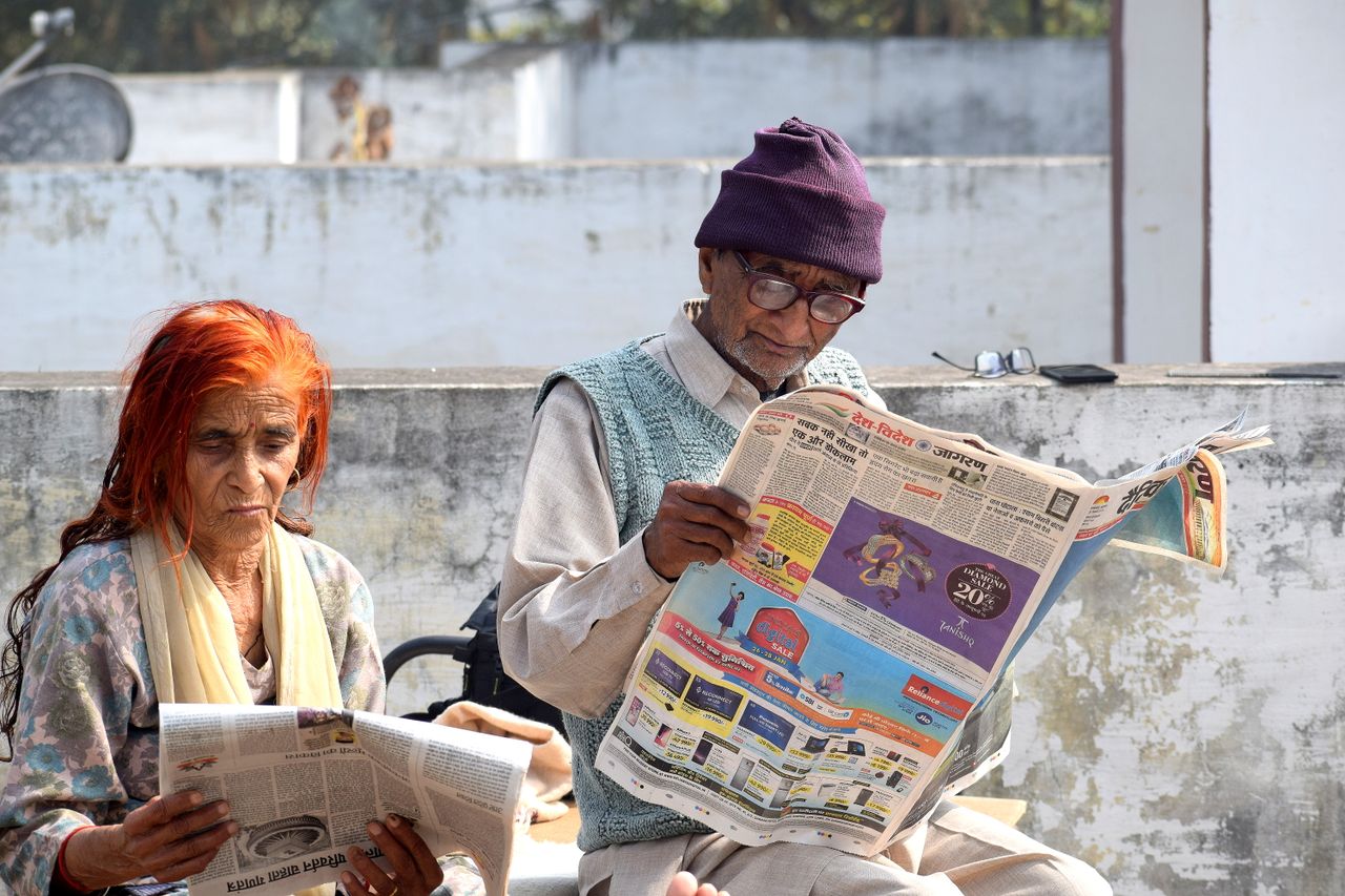 gallery-6 An elderly couple enjoys reading newspapers outside on a sunny day, reflecting a peaceful moment.