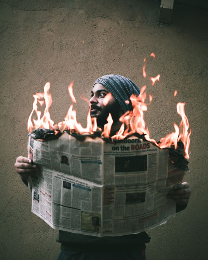 gallery-4 Unshaven ethnic male in knitted hat burning newspaper with bright flame while standing near concrete wall and looking away