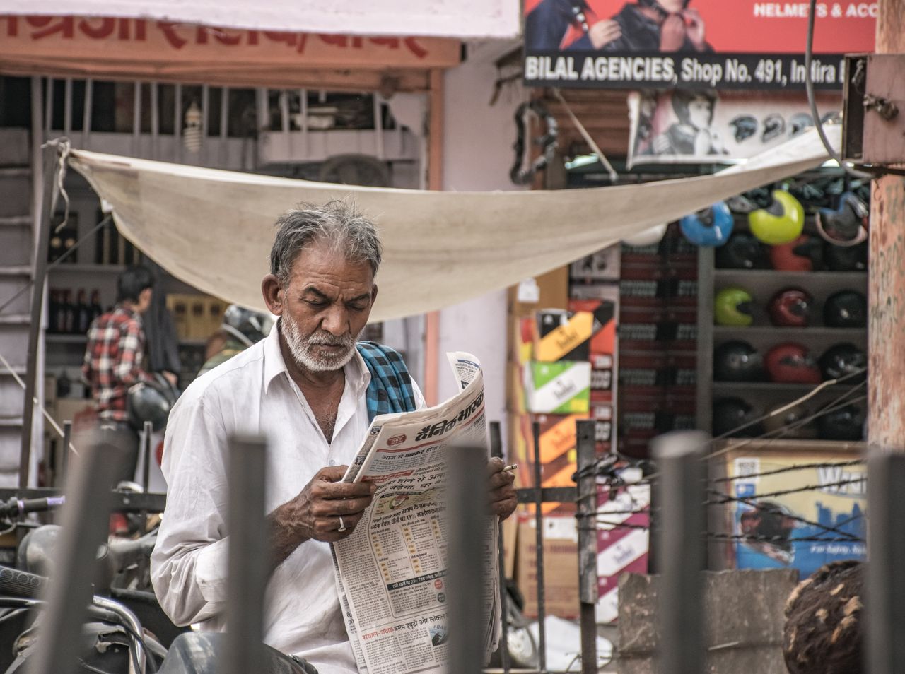 contact-img Elderly man immersed in a newspaper amidst a bustling urban street market.